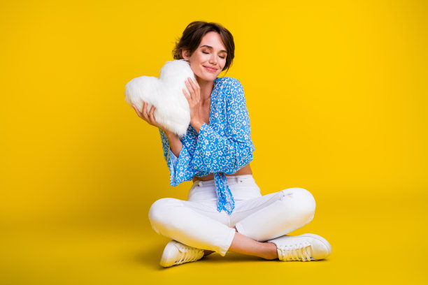 满意的温柔女人的全尺寸照片，穿着蓝色上衣，坐在那里，在黄色背景上的颧骨上抱着蓬松的枕头图片下载