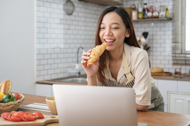 在现代厨房里吃早餐时吃羊角面包的快乐女人的照片。图片下载