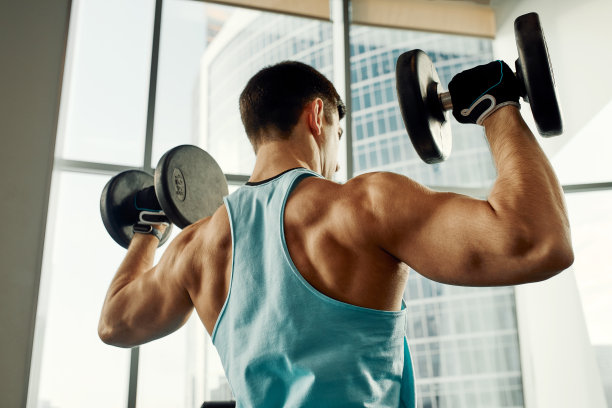 一个男人倚在健身房的运动板凳上拿着哑铃做运动。灰色背景下，一个穿着运动服、体格健壮的性感肌肉男人的照片。力量和动力，运动，健身目标图片下载