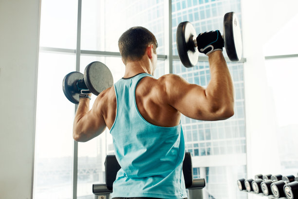 一个男人倚在健身房的运动板凳上拿着哑铃做运动。灰色背景下，一个穿着运动服、体格健壮的性感肌肉男人的照片。力量和动力，运动，健身目标图片下载