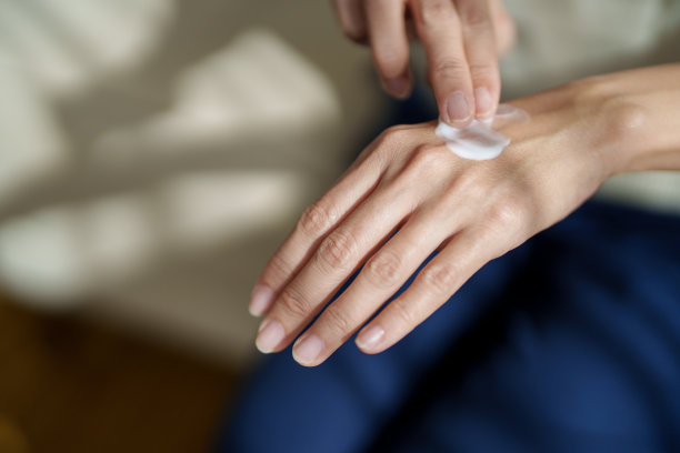 在这张剪短的图片中，一位中年亚洲女性正在用润肤霜涂抹双手干燥的皮肤，阳光温柔地照在她的皮肤上，突显出滋养的姿态。图片下载