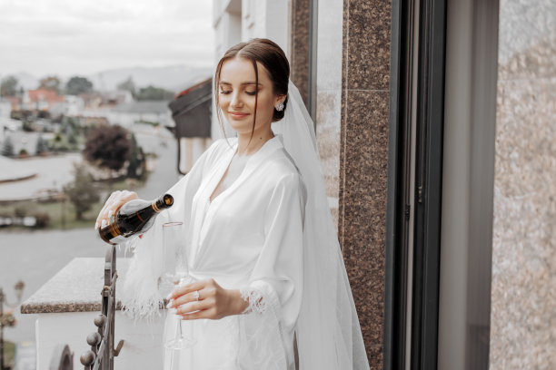 一个美丽的新娘，头发上戴着长长的面纱，穿着优雅的白色睡衣，站在阳台上俯瞰城市，在婚礼的早晨喝着饮料。图片下载