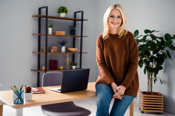 照片满意的金发退休妇女坐在台式机上网本放松在她的家庭办公室室内阁楼设计室内图片下载