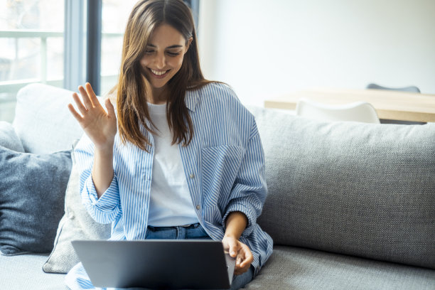 积极开朗的女孩全身照片坐在沙发上盘腿用电脑视频打电话给她的朋友在家里的室内。千禧一代女性在沙发上使用笔记本电脑在室内休闲图片下载