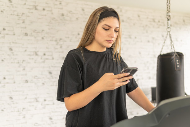 年轻的拉丁女子在健身休息时在跑步机上看她的手机图片下载