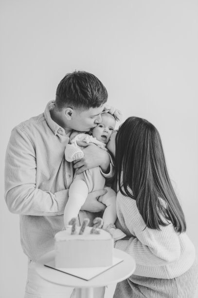 家庭庆祝六个月大的婴儿生日聚会。黑白照片。爸爸妈妈亲吻隔离在墙上的可爱女孩。爸爸、妈妈牵着手逗小孩。创意生日蛋糕与蜡烛。图片下载