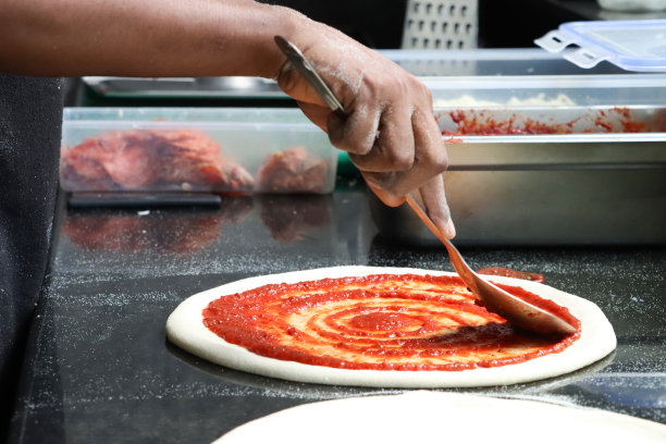 特写照片中，无法辨认的Pizzaiolo(披萨厨师)用金属勺子在未煮熟的披萨面团上涂上浓郁的番茄沙司，商业厨房背景，焦点在前景图片下载