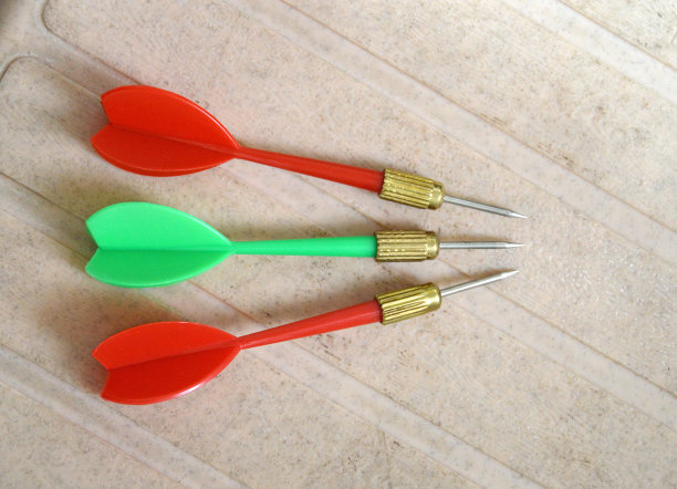 三个飞镖的水平图片，两个红色和一个绿色的尖箭头与尖锐的金属头与尖的尖端排列在米色木制纹理瓷砖背景与复制空间图片下载
