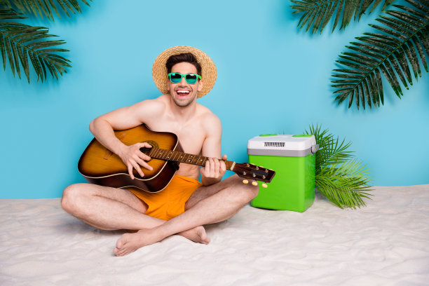 全长照片无忧无虑可爱的家伙泳装太阳帽坐在沙滩上弹吉他孤立的蓝色背景图片下载