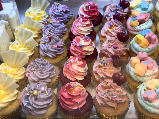 全画幅图片，一排排的纸杯蛋糕在银纸蛋糕盒装饰与糖霜漩涡在玻璃镶板面包店货架展示，抬高视图图片下载