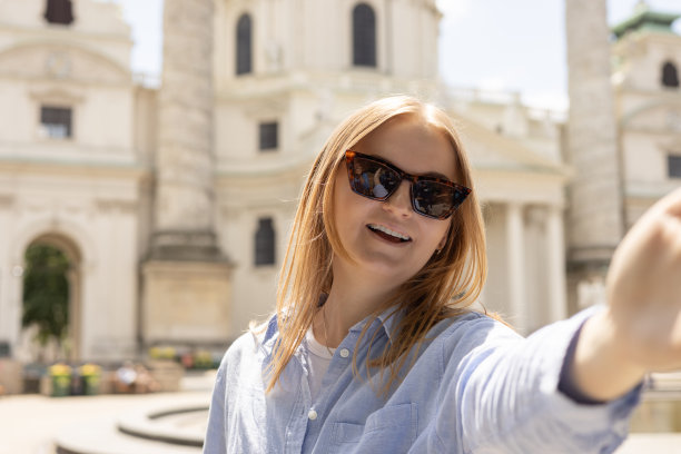 照片轻松迷人积极的年轻女子拿着相机，在市中心户外自拍夏天。旅游度假理念的生活方式。观点。维也纳圣查尔斯教堂图片下载