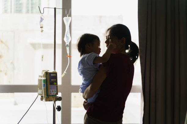 后视图剪影照片，亚洲母亲在医院康复时抱着并安慰她的儿子，手里拿着静脉点滴。可爱的亚洲孩子感染病毒住院了。婴儿保健医疗理念。图片下载