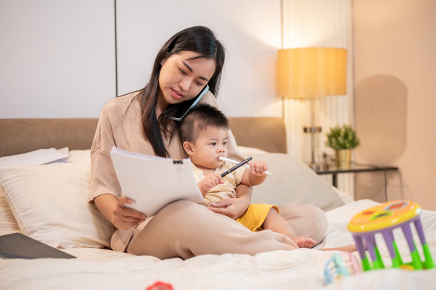 一位忙碌的亚洲妈妈抱着她的儿子，一边打电话一边在床上处理文件。图片下载