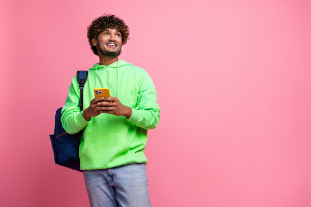 照片中的年轻人穿着运动衫后面背着帆布背包，浏览手机，看学生的模拟价目表，孤立在粉红色的背景上图片下载