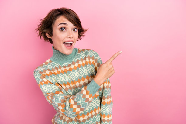 照片中美丽的年轻女子穿着秋天的印花套头衫指着空白的空间，哇哦，omg提供孤立的粉红色背景图片下载