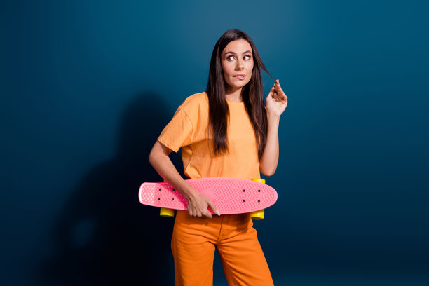 漂亮女人的照片咬嘴唇，手指触摸黑发卷发持有粉红色的长板看起来模型孤立在深蓝色的背景图片下载