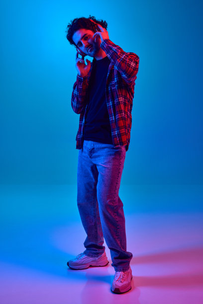 年轻人的全身凝胶肖像，卷发，没有剃光的脸，戴着耳机听音乐在霓虹灯蓝色背景图片下载