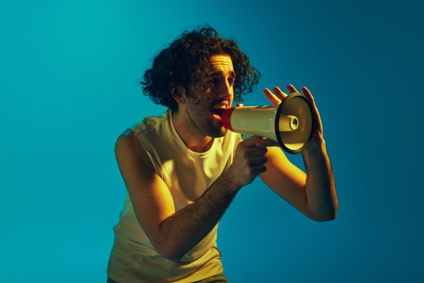 一个情绪化的卷发年轻人，在霓虹灯的蓝色背景下，用扩音器大声喊叫。新闻、公告图片下载