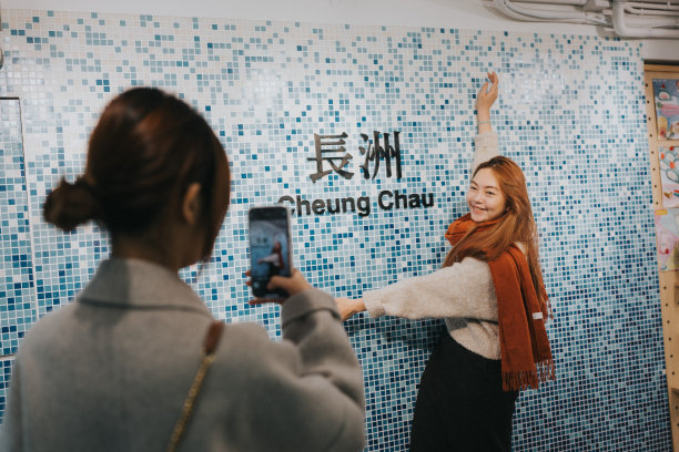 在这幅图片中，一名男子拍摄了一名微笑的女子站在马赛克图案和中英文“长洲”字样前的照片。图片下载