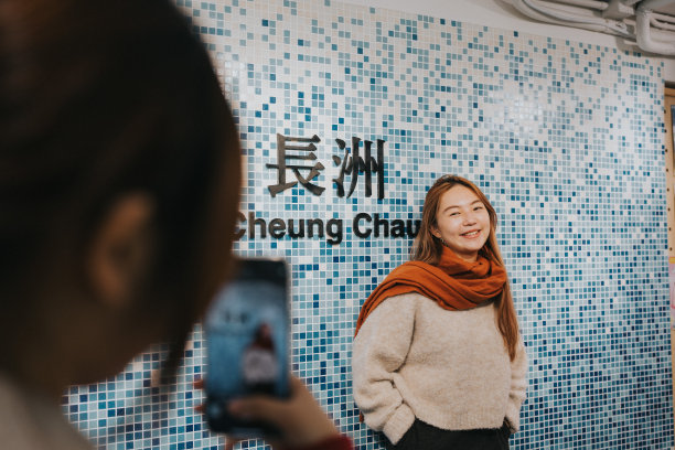 在这幅图片中，一名男子拍摄了一名微笑的女子站在马赛克图案和中英文“长洲”字样前的照片。图片下载