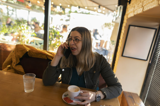 快乐的年轻女子一边用手机一边在咖啡店喝咖啡。戴着圆眼镜的黑发女郎拍下了真实的电话交谈，真情流露。图片下载