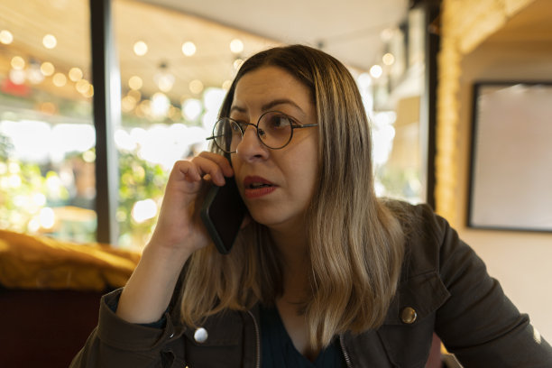 快乐的年轻女子一边用手机一边在咖啡店喝咖啡。戴着圆眼镜的黑发女郎拍下了真实的电话交谈，真情流露。图片下载