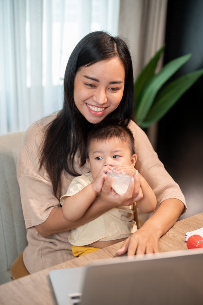 一位乐观的亚洲母亲一边用笔记本电脑工作，一边用奶瓶喂她的男婴。图片下载