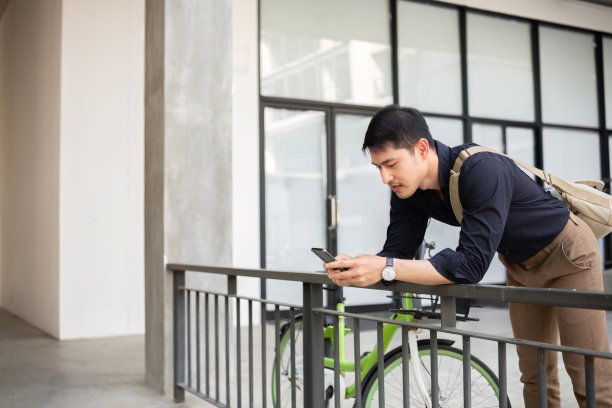 一位年轻的亚洲商人骑着自行车去上班。站在办公室外面打电话，谈生意。减少能源和减少空气污染的理念。图片下载