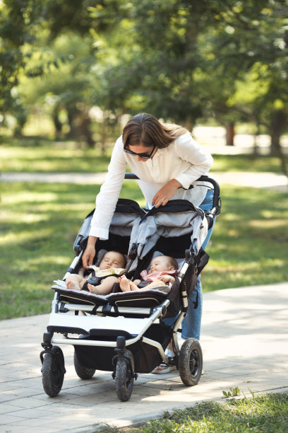 垂直照片生活方式年轻随意的白人母亲照顾两个小双胞胎女孩在一个双人婴儿车，在城市公园散步图片下载