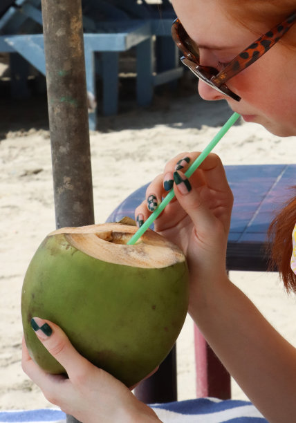 迷人的特写图片，红发长发的女人在阳光明媚的日子里戴着太阳镜，坐在沙滩伞遮阳伞下，用吸管拿着椰子，沙滩背景，侧面视图，重点在前景图片下载