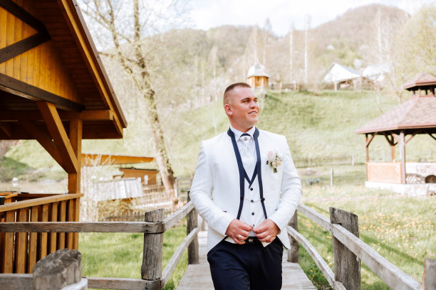 新郎穿着白色夹克和黑色裤子，微笑着摆姿势，扣上衬衫上的纽扣。婚礼的肖像。图片下载