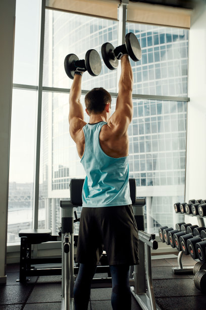 一个男人倚在健身房的运动板凳上拿着哑铃做运动。灰色背景下，一个穿着运动服、体格健壮的性感肌肉男人的照片。力量和动力，运动，健身目标图片下载
