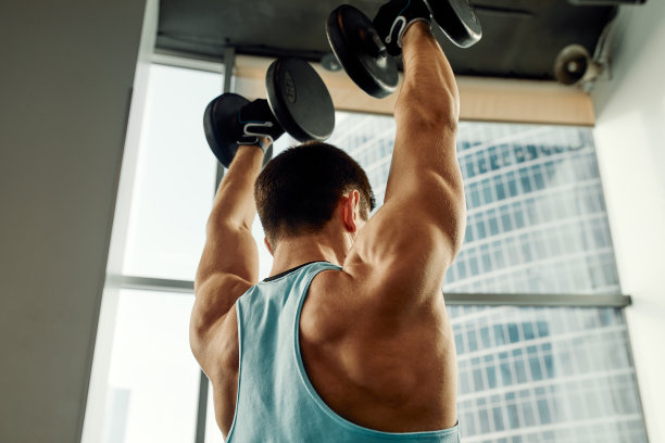 一个男人倚在健身房的运动板凳上拿着哑铃做运动。灰色背景下，一个穿着运动服、体格健壮的性感肌肉男人的照片。力量和动力，运动，健身目标图片下载