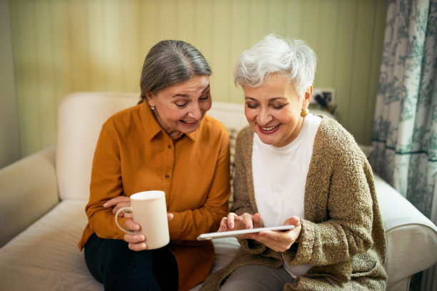 两个优雅快乐的退休高加索女性在室内放松，喝咖啡，在便携式电子设备上上网，浏览表情包，有趣的图片或通过社交媒体在线观看视频图片下载