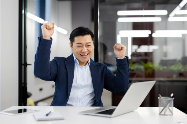 快乐的男人在工作场所跳舞，成功的亚洲商人穿着西装坐在工作场所，在办公室里拿着笔记本电脑，庆祝成功的成果，微笑着满足图片下载