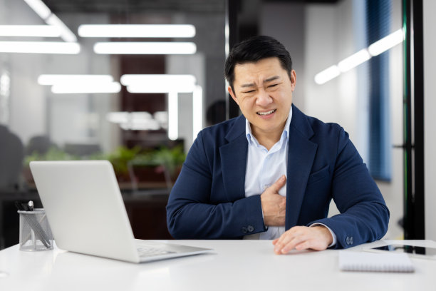 心酸的商人，胸痛的亚洲人在办公室里的工作场所，成熟的老板穿着西装，手放在胸前，坐在桌子上用笔记本电脑图片下载