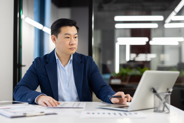 办公室里的文书工作背后是认真思考的人，西装革履的成熟亚洲人在表格里填写数据，用笔写字，办公室里的商人老板用笔记本电脑图片下载