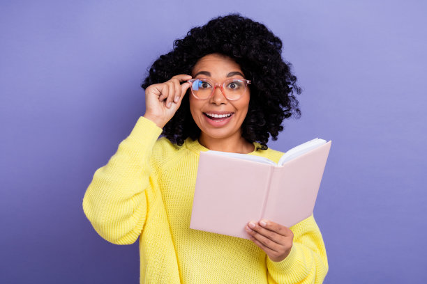 照片聪明的女人与非洲发型穿着黄色毛衣触摸眼镜拿书孤立的紫色背景图片下载