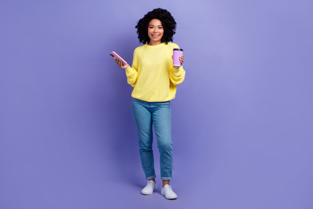 开朗可爱的人的全身照片，穿着针织套头衫，拿着智能手机外卖咖啡杯，孤立在紫色的背景上图片下载