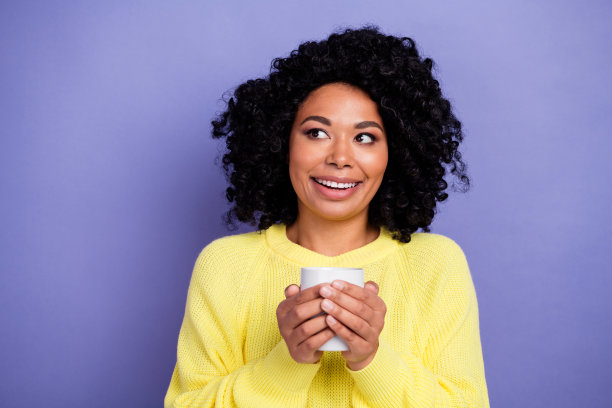照片中满意的女人留着非洲式发型，穿着黄色毛衣，拿着热杯子，看着紫色背景上的空白空间图片下载