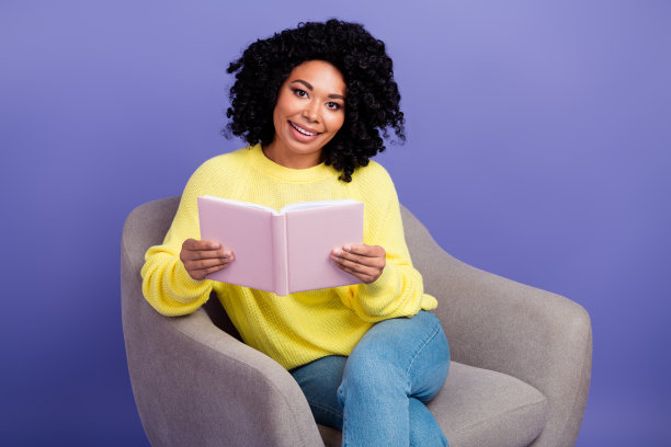 照片中聪明开朗的女孩穿着黄色的羽绒服，坐在扶手椅上，拿着紫色背景上孤立的书图片下载