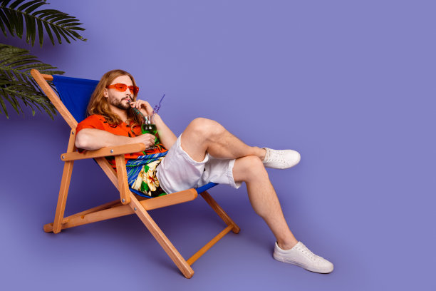 满意的男人穿着印花衬衫的全身照片躺在躺椅上喝鸡尾酒在沙滩上孤立的紫色背景图片下载