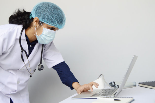 一个女人或女士或女医生戴着外科口罩，戴着帽子，穿着白色的实验室外套，脖子上挂着听诊器，站着，弯腰工作，在灰色背景上聚焦一台笔记本电脑，上面有文字图片的复制空间图片下载