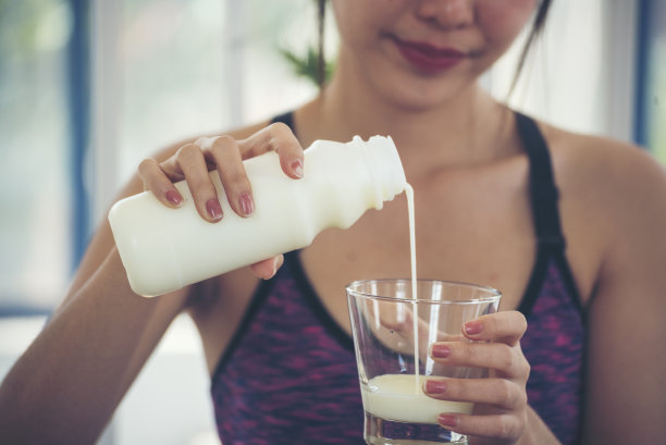 女人的手拿着白色的玻璃杯，牛奶从瓶子里倒出来。亚洲女性笑着笑着看镜头保健居家健身生活方式。美女饮用新鲜牛奶，低脂，无乳糖图片下载