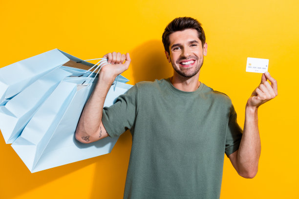 照片积极聪明的男子鬃毛穿着卡其布t恤拿着新衣服的袋子信用卡孤立的黄色背景图片下载