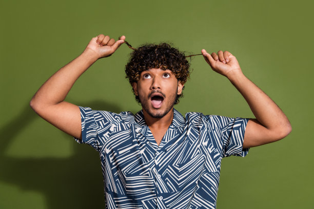 照片中震惊的滑稽男子穿着印花衬衫，拿着头发卷发孤立的绿色背景图片下载