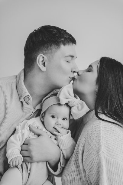 父亲亲吻母亲拥抱女儿。幸福的家庭在节日里拥抱可爱的宝宝。爸爸，妈妈牵着婴儿的手。爸爸，妈妈抱着小女孩，头发上扎着蝴蝶结，孤立在墙上。黑白照片。家庭。图片下载