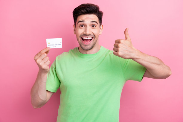 照片中，兴奋的男子穿着绿色t恤竖起大拇指，就像拿着塑料卡，广告信用系统孤立在粉红色的背景上图片下载