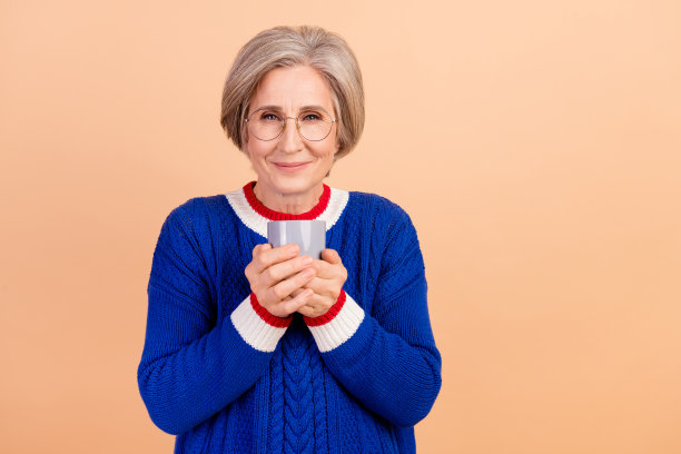 照片中可爱可爱的退休妇女穿着时髦的蓝色衣服，享受着米色背景上的美味芳香饮料图片下载