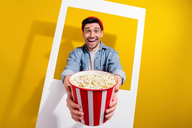 照片心情愉快的男人穿着牛仔裤衬衫邀请你吃爆米花钉相框孤立的黄色背景图片下载